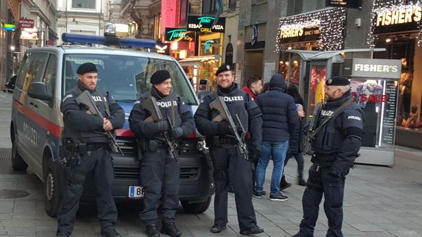 Vier bewaffnete Polizisten stehen vor einem Polizeiwagen in einer belebten Straße.