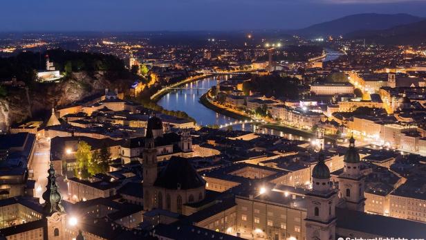 Nachtsicht auf Salzburg mit der Salzach, beleuchteten Gebäuden und Bergen im Hintergrund.