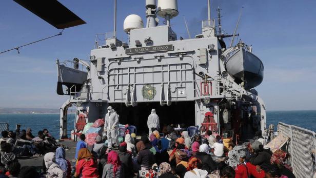 Viele Menschen sitzen auf dem Deck eines Schiffes, einige in Schutzanzügen.