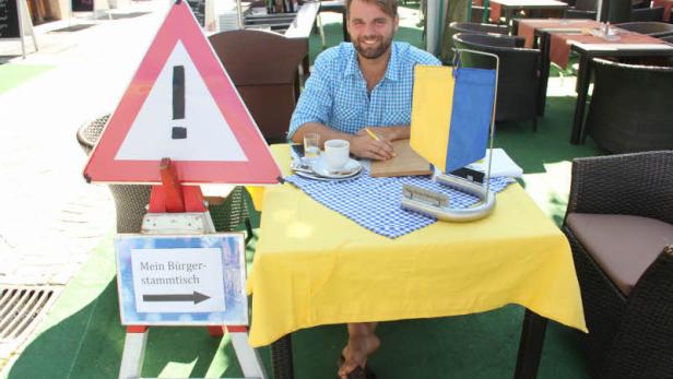 Ein Mann sitzt barfuß an einem Tisch mit einem Schild „Mein Bürgerstammtisch“.