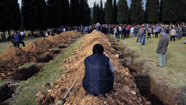 Eine Person sitzt auf einem Erdhügel vor einer Reihe offener Gräber, während sich im Hintergrund eine Menschenmenge versammelt.