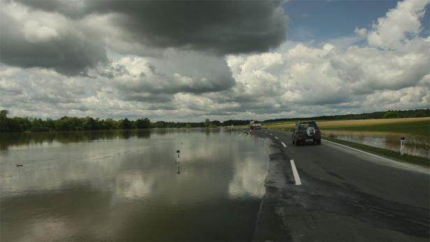 Eine überflutete Straße mit einem Auto und einem Lastwagen unter bewölktem Himmel.