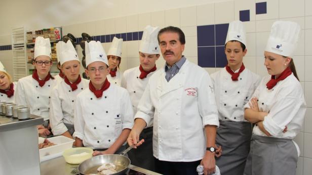 Ein Koch mit Schnurrbart unterrichtet eine Gruppe junger Köche in einer Küche.