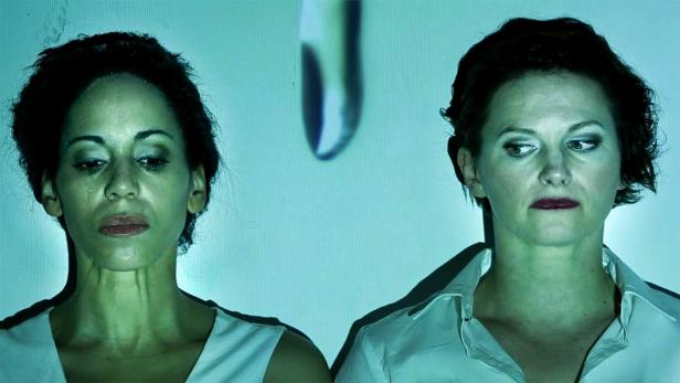 Zwei Frauen stehen vor einer hellblauen Wand, beleuchtet von einem Licht.