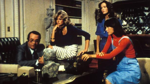 Ein Mann in einem Anzug sitzt an einem Schreibtisch, umgeben von drei Frauen.