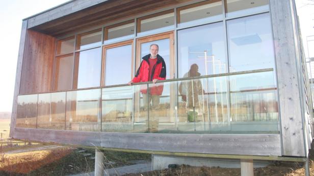 Ein Mann in roter Jacke steht auf dem Balkon eines modernen Holzhauses.