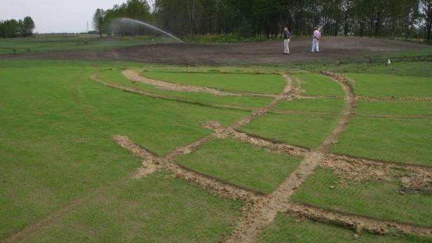 Ein Rasenfeld mit Bewässerungssystem und zwei Personen im Hintergrund.