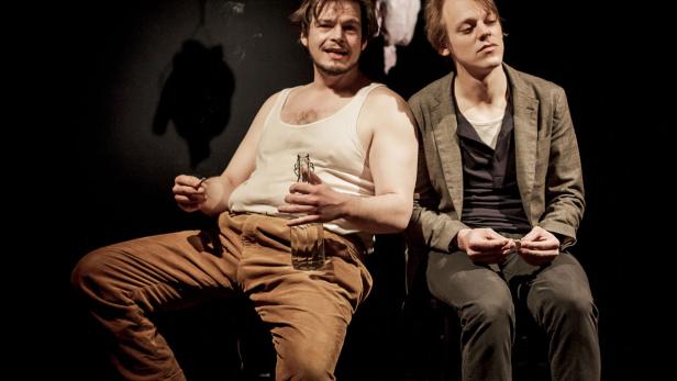 Zwei Männer sitzen auf Stühlen, einer mit Unterhemd und Flasche, der andere mit Jackett.