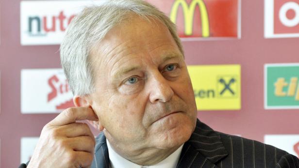 Ein Mann im Anzug kratzt sich am Ohr vor einem Hintergrund mit Werbelogos.