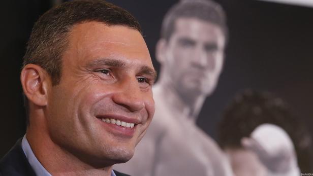 Wladimir Klitschko lächelt vor einem verschwommenen Boxporträt.