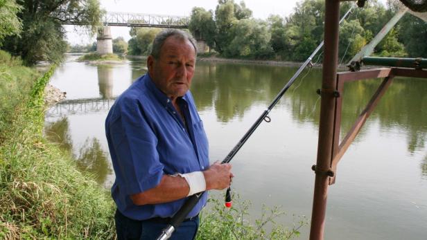 Ein Mann mit bandagiertem Handgelenk steht am Flussufer und hält eine Angelrute.