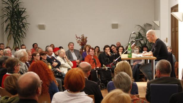 Ein Mann liest vor Publikum aus einem Buch vor.