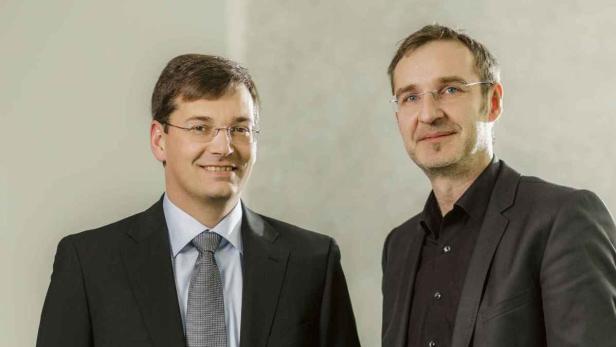 Zwei Männer in Anzügen stehen nebeneinander und lächeln.