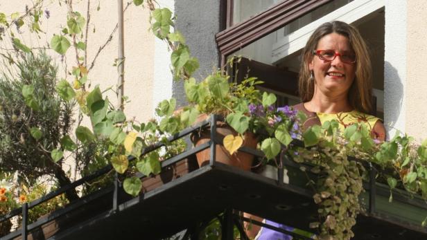 Eine Frau mit roter Brille schaut von einem begrünten Balkon.