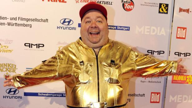 Ein Mann mit roter Kappe und goldener Jacke steht vor einer Wand mit Logos.