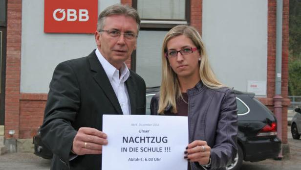 Zwei Personen halten ein Schild mit der Aufschrift „Unser Nachtzug in die Schule!!! Abfahrt: 6.03 Uhr“ vor einem ÖBB-Gebäude.