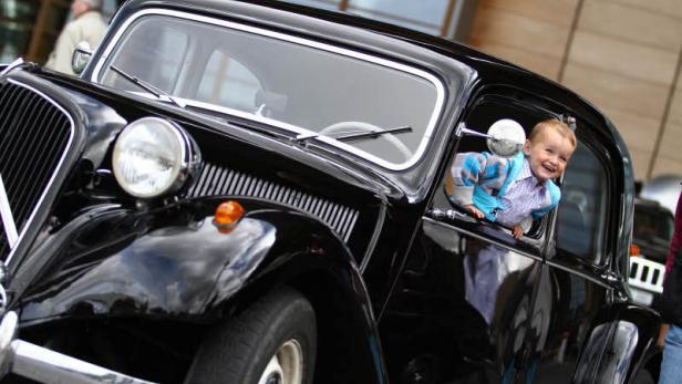 Ein lachender Junge lehnt aus dem Fenster eines schwarzen Oldtimers.