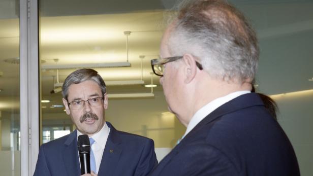 Ein Mann mit Brille und Schnurrbart spricht in ein Mikrofon, während ein anderer Mann ihm zuhört.