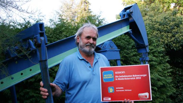 Ein Mann hält ein Schild mit der Aufschrift „Gesucht: Die beliebtesten Nahversorger in Niederösterreich 2012“ vor einer blauen Förderanlage.