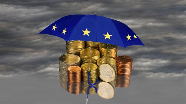 Ein Regenschirm mit EU-Flagge schützt einen Stapel von Euromünzen vor einem dunklen Himmel.