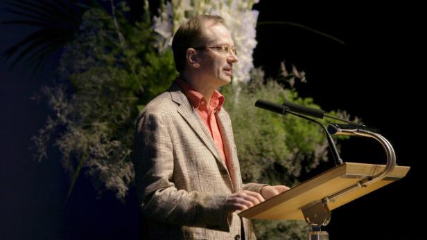 Ein Mann mit Brille hält eine Rede an einem Rednerpult vor floralem Hintergrund.