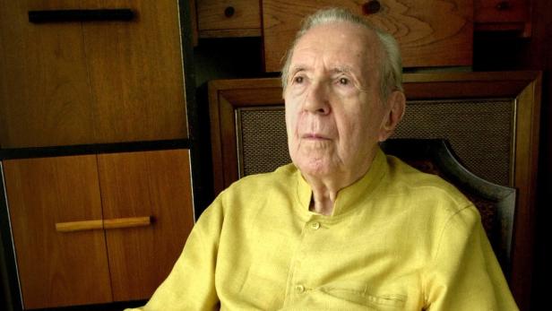 Ein älterer Mann in einem gelben Hemd sitzt vor einem Holzschrank.