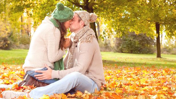 Ein Paar in Strickmützen küsst sich inmitten von Herbstlaub.