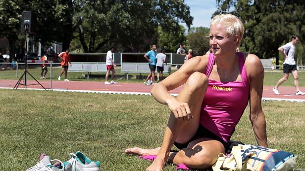Eine blonde Sportlerin sitzt auf einer Wiese neben einer Laufbahn.