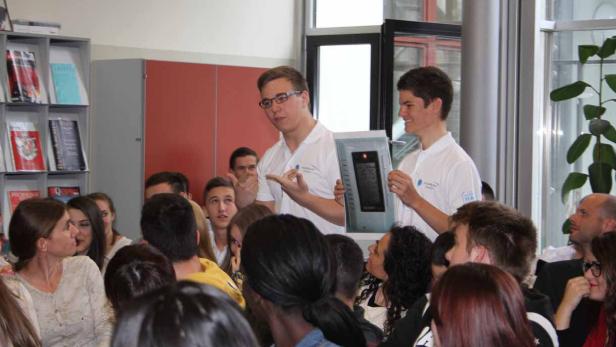 Zwei junge Männer präsentieren ein Gerät vor einer Gruppe von Zuhörern.