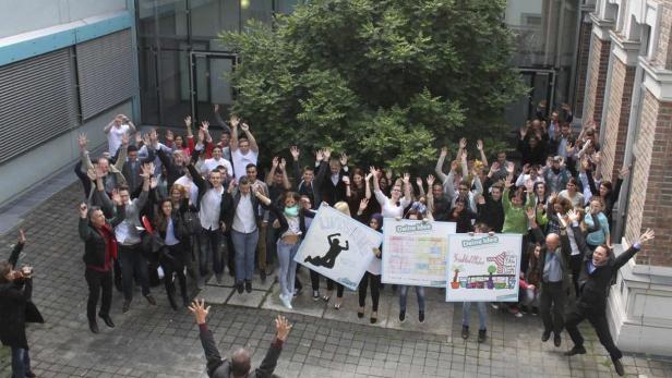 Eine Gruppe von Menschen springt mit erhobenen Händen vor einem Gebäude in die Luft.