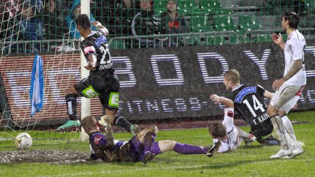 Ein Fußballspieler schießt den Ball ins Tor, während der Torwart im Schlamm liegt.