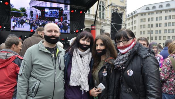 Eine Gruppe von Menschen mit gestrickten Bärten steht vor einer Bühne.
