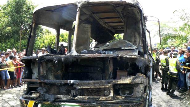 Ein ausgebrannter Bus steht vor einer Menschenmenge und Sicherheitskräften.