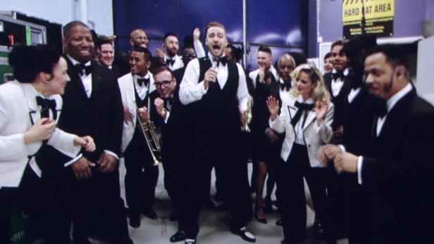 Justin Timberlake singt mit einem Mikrofon, umgeben von einer Band in Anzügen.