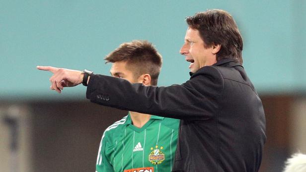 Ein Trainer gestikuliert und gibt Anweisungen, während ein Fußballspieler im Hintergrund steht.