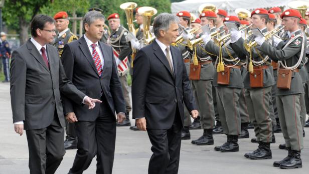 Drei Männer in Anzügen gehen vor einer Militärkapelle her.