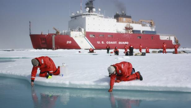 Zwei Personen in roten Anzügen nehmen Wasserproben auf einer Eisscholle vor einem Schiff der US-Küstenwache.