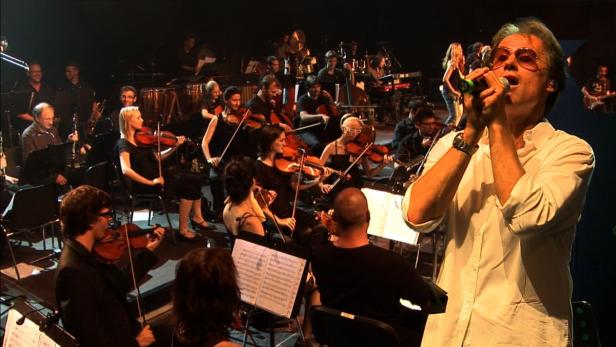 Ein Sänger tritt mit einem Orchester im Hintergrund auf einer Bühne auf.