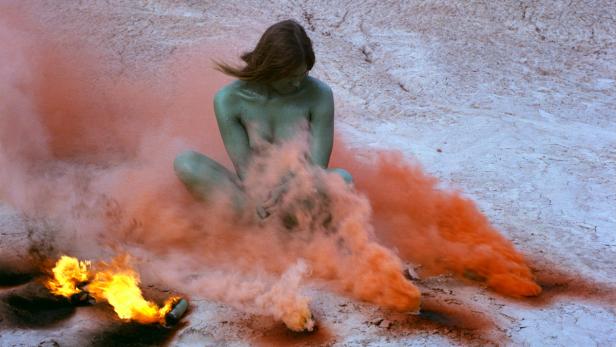 Eine Frau sitzt inmitten von Rauch und Feuer in einer kargen Landschaft.