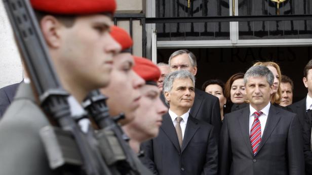 Soldaten in roter Barett stehen vor einem Gebäude, dahinter eine Gruppe von Personen in Anzügen.