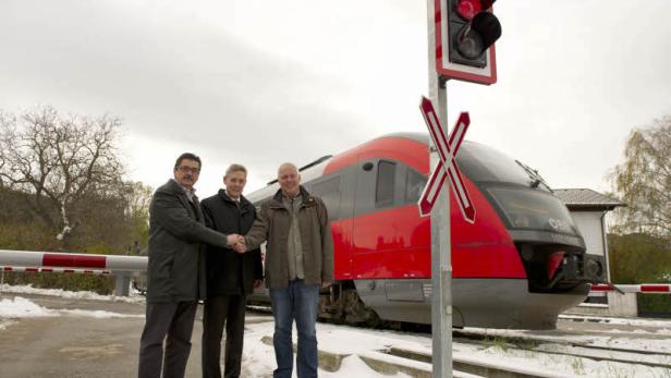 Drei Männer schütteln sich vor einem roten ÖBB-Zug an einem Bahnübergang die Hände.