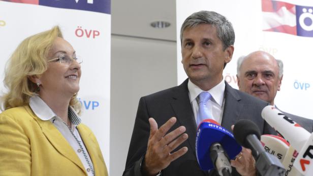 Ein Mann in Anzug spricht vor Mikrofonen, flankiert von zwei weiteren Personen vor einem ÖVP-Banner.