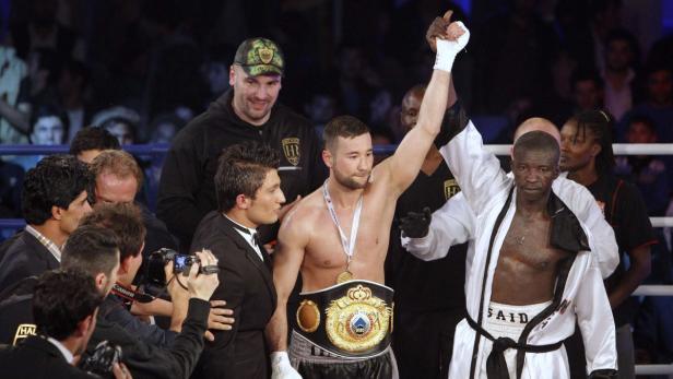 Ein Boxer mit Siegergürtel wird von seinem Trainer gefeiert, während sein Gegner daneben steht.