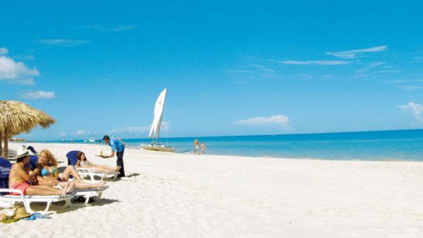 Ein Kellner serviert Getränke an einem sonnigen Strand mit Liegestühlen und einem Segelboot im Hintergrund.