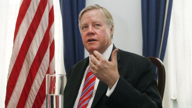 Ein Mann in Anzug und Krawatte gestikuliert vor einer amerikanischen Flagge.