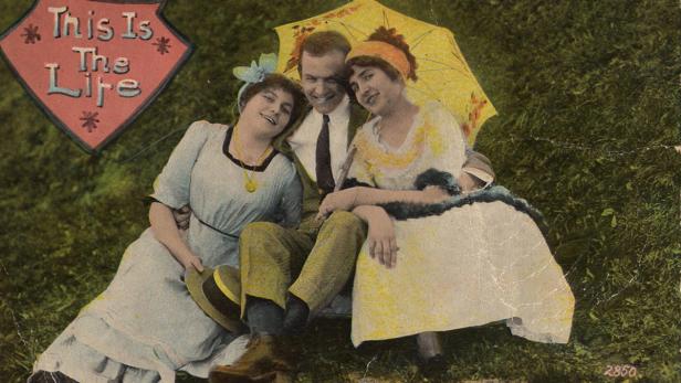 Ein Mann sitzt mit zwei Frauen im Gras unter einem Sonnenschirm.