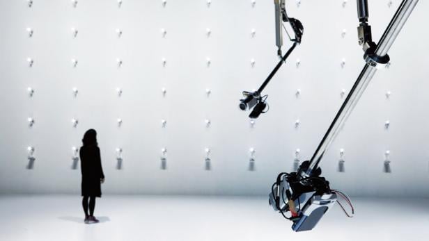 Eine Frau steht in einem weißen Raum vor einer Wand mit Lichtern und Roboterkameras.