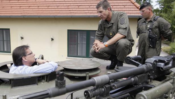 Ein Mann in Zivil sitzt in einem Panzer, während zwei Soldaten ihn betrachten.