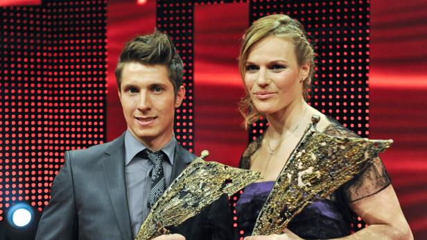 Ein Mann im Anzug und eine Frau halten goldene Trophäen vor einem roten Hintergrund.