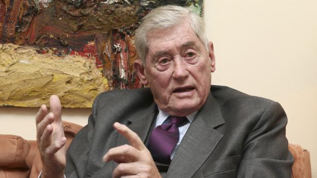 Ein älterer Mann in einem grauen Anzug gestikuliert vor einem abstrakten Gemälde.